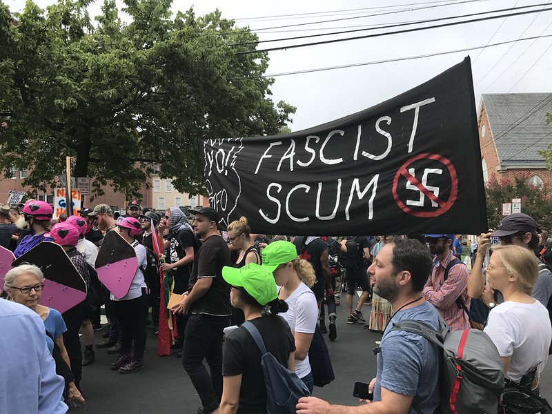 A sizable crowd is gathered outside Emancipation Park (now called Market Street Park and was Robert E. Lee Park until 2017) with a couple of individuals in the middle holding a large black sign that reads “Fascist Scum” in large white letters with a crossed out swastika symbol in the bottom right corner of the sign. Two individuals wearing green hats in the front are legal observers.