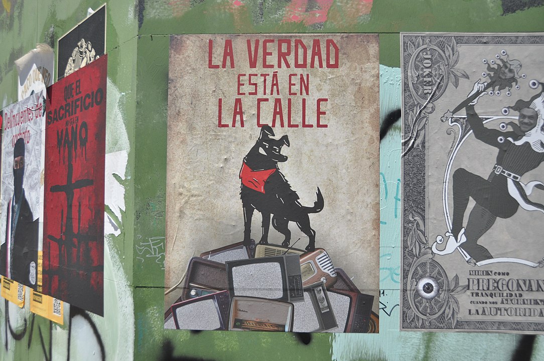 Photo of a series of protest posters on outdoor wall, from Chilean uprising in 2019. The central poster shows a black dog standing on a pile of broken televisions, with the slogan in spanish, “The truth is in the streets.”