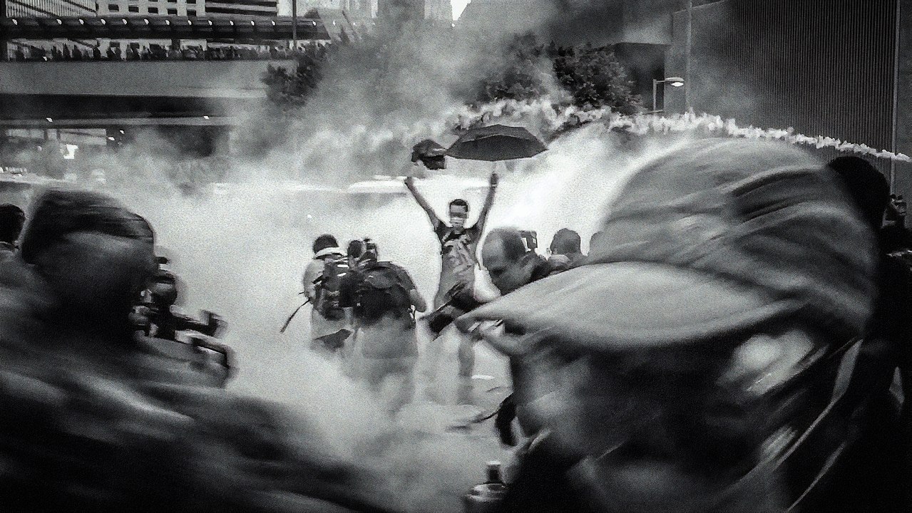 Lone protester holding up an umbrella, in the midst of chaos, tear gas, and massive protests, and protestors in gas masks, during the 2014 Umbrella Movement uprising in Hong Kong.