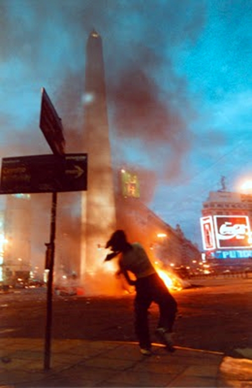 Image of youth in foreground, apparently throwing a petrol bomb, with the burning street, and fire in front of the obelisk in downtown Buenos Aires, Argentina, December 2001.