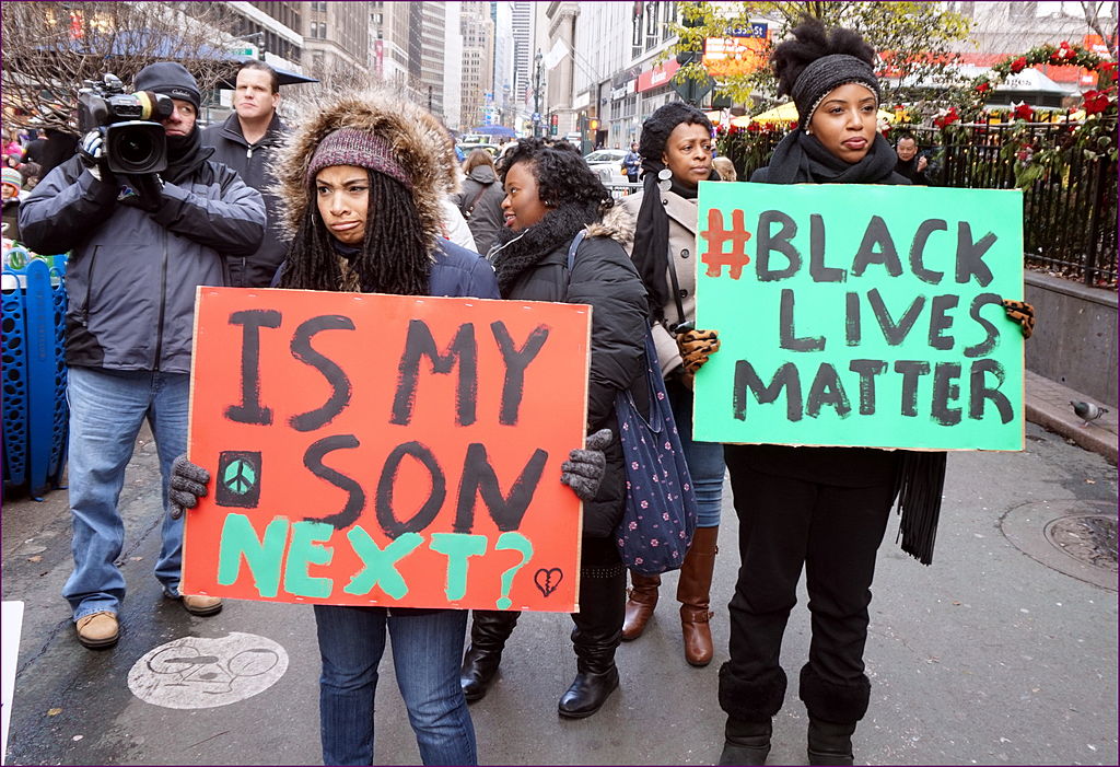 Several activists in outdoor clothing are gathered on an urban street holding protest signs reading "Is my son next?" and "#BlackLiveMatter"