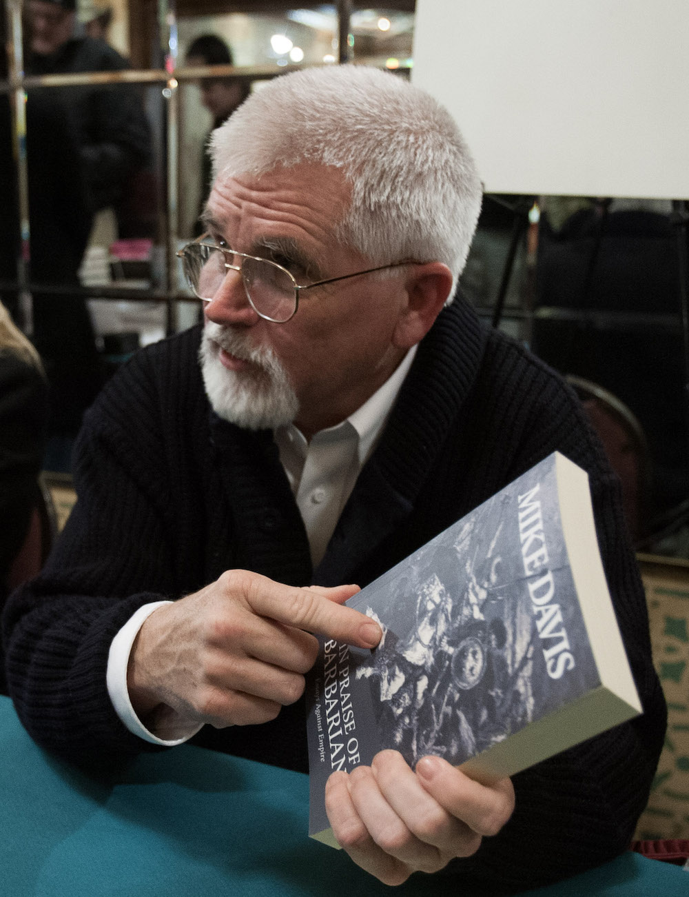 Author Mike Davis, a white man with white hair, beard, and glasses, holds his book In Praise of Barbarians.