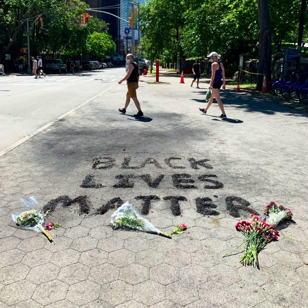 The words Black Lives Matter are spelled out in ash on a sidewalk, with the word Matter partially covered by three bouquets of flowers. Photo by @jameswest2020.