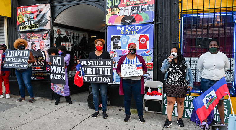 A line of demonstrators stand in a line along a sidewalk holding Haitian flags and protest signs that read TRUTH DEMANDS ACCOUNTABILITY, NO MORE RACISM, JUSTICE IS TRUTH IN ACTION, and Halt Deportations of Haitian Asylum Seekers @ The Border.
