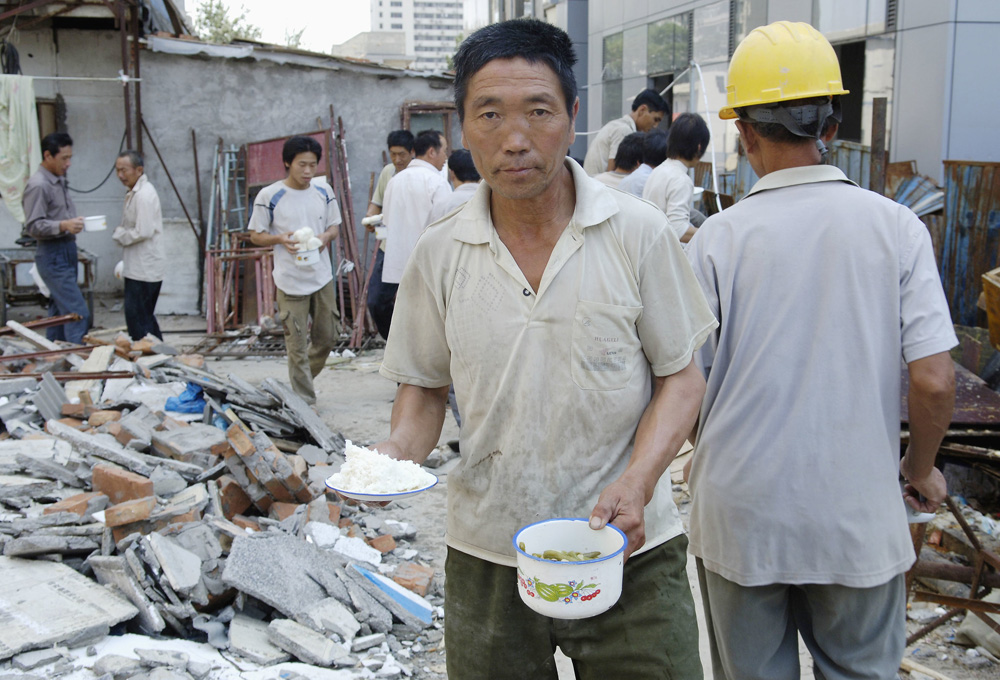 Image of a Chinese worker with several others amid construction debris, holding bowls of green beans and rice