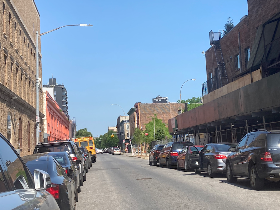 Photograph of Abermale Road in Brooklyn lined with buildings and cars on either side stretches beneath a cloudless blue sky, facing Bedford Avenue in the distance. 