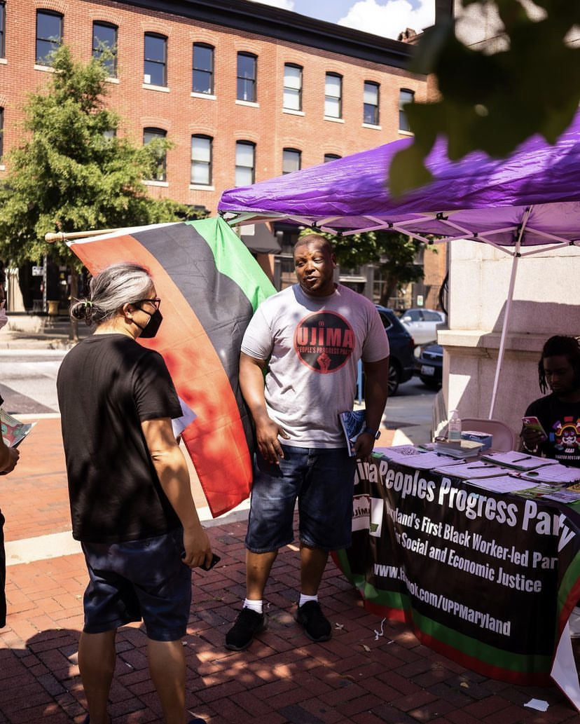 A man wearing a T-shirt featuring a red fist and the logo for the Ujima People’s Progress Party speaks with a masked individual next to a table with a large green, black, and red Pan-African Flag and a banner that reads, “Ujima People’s Progress Party, Maryland’s First Black Worker-Led Party for Social and Economic Justice.”