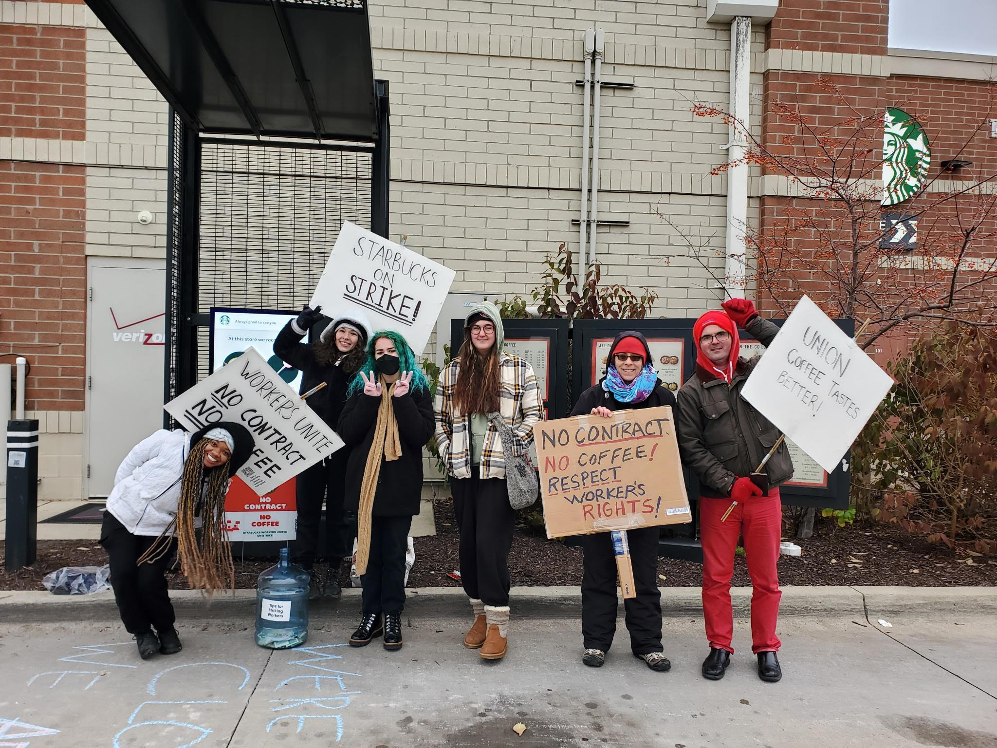Six young Starbucks workers in cold-weather garb hold signs reading Starbucks on Strike! No Contract No Coffee! and Respect Workers' Rights!