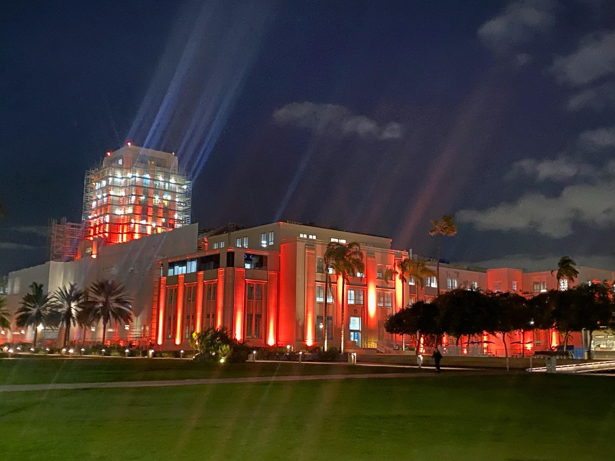 The San Diego County Administration Building, a large rectangular building, is shown beneath a partly cloudy night sky and is lit with bright neon orange lights across the building’s façade.