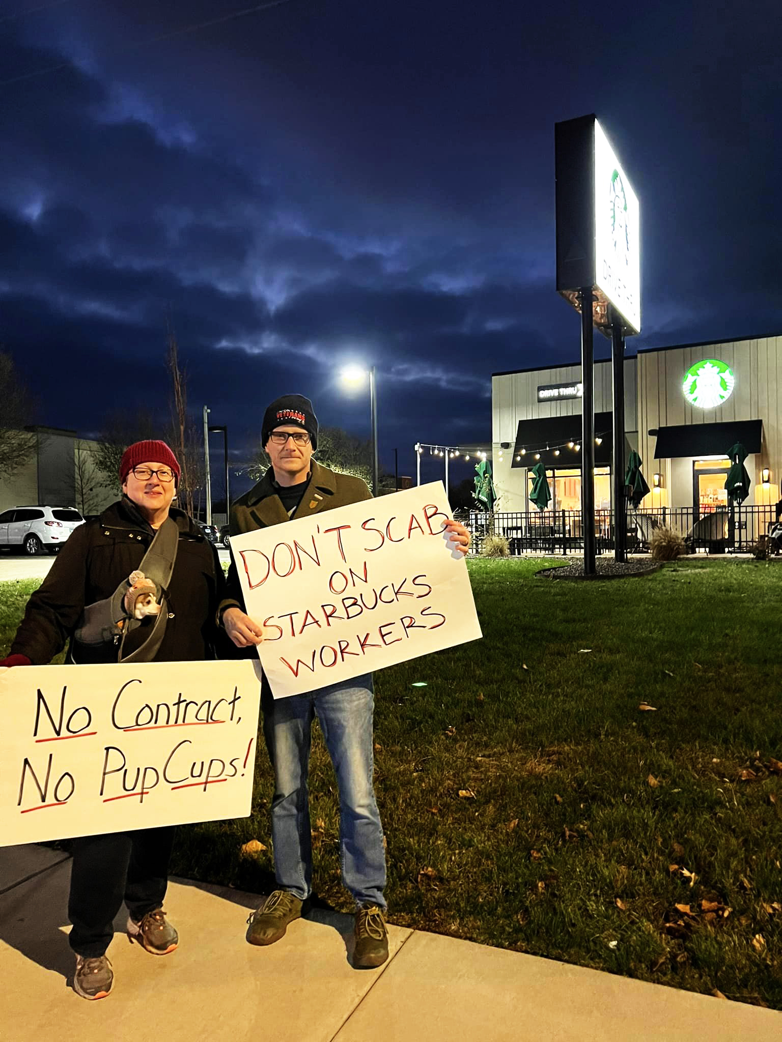 Two picketers, including one with a small dog in a strapon carrier, hold signs that read “No Contract No Pup Cups!” and “DON’T SCAB ON STARBUCKS WORKERS.”