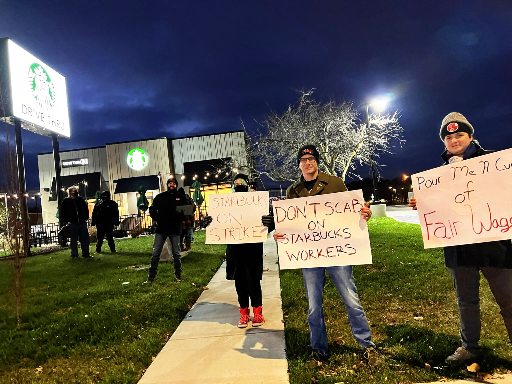 Picketers stand scattered across the lawn outside a Starbucks drive-through. One of their signs reads, “Pour Me A Cup of Fair Wages.”