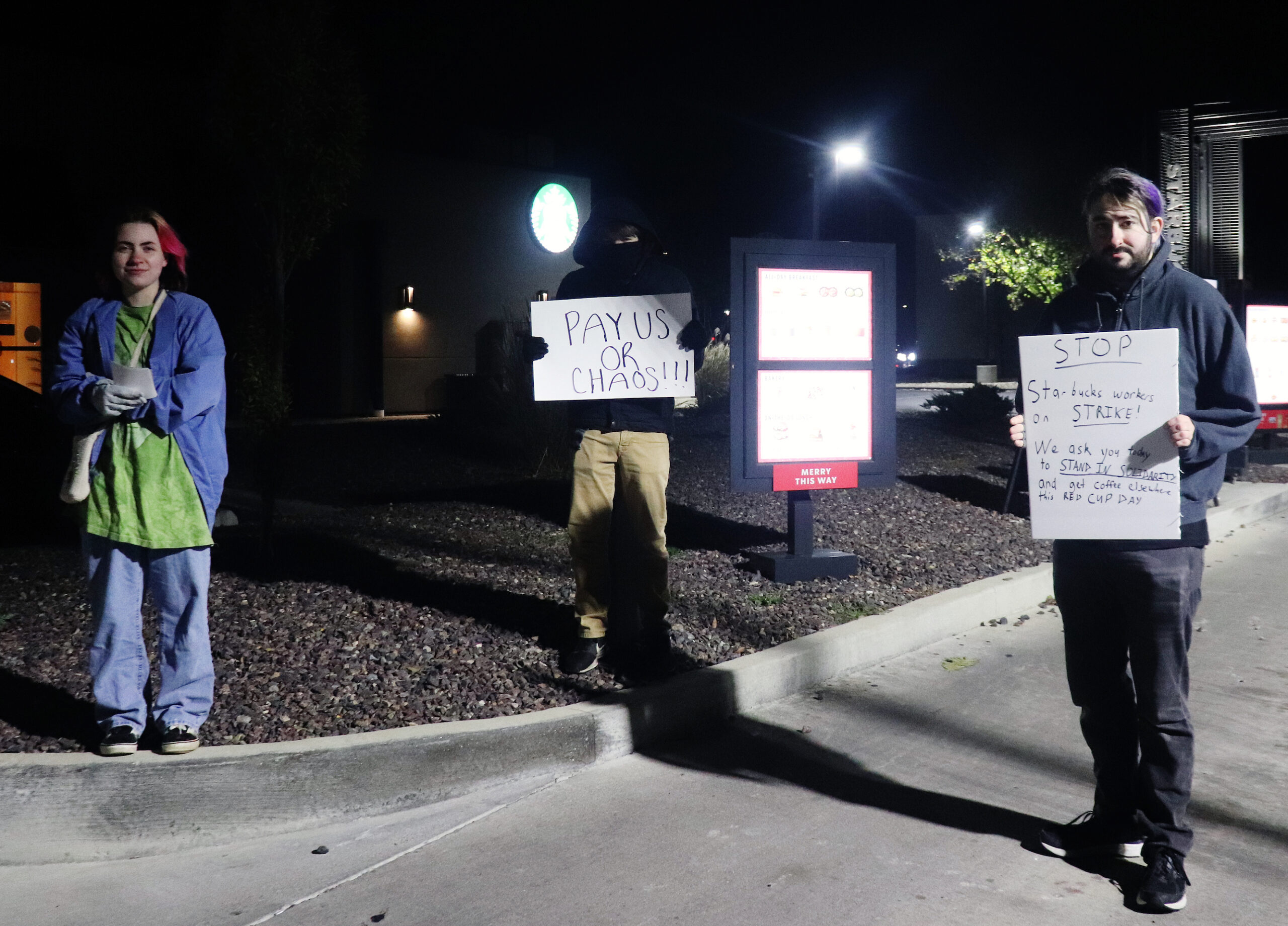 Workers stand in entrance to the Starbucks drive-through. One holds a sign that reads, “PAY US OR CHAOS!!!” A second implores customers not to cross the picket line.