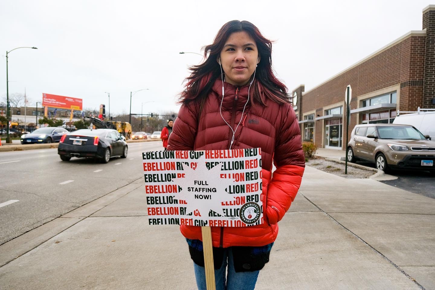 A worker with straight red dyed hair in a red puffer jacket holds a picket sign that reads, “FULL STAFFING NOW RED CUP REBELLION.”Link: http://www.loveandstrugglephotos.com/