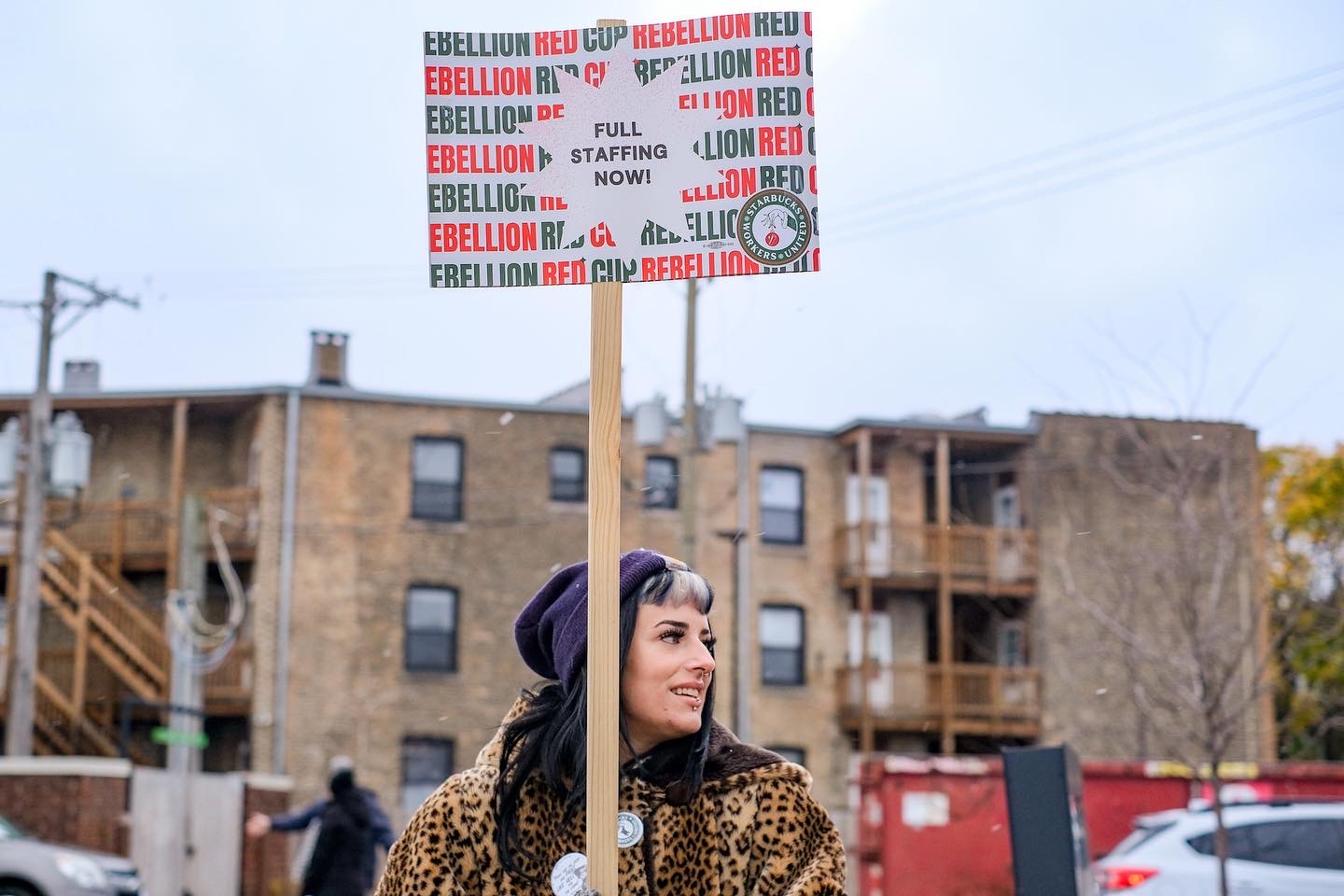 A worker with streaked hair and bangs wearing a leopard print jacket holds a sign high above their head that reads, “FULL STAFFING NOW.”