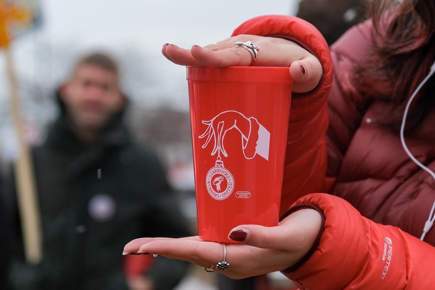 A pair of hands holds a plastic red cup emblazoned with a cartoon hand holding a Christmas ornament that features the Starbucks Workers United union logo.