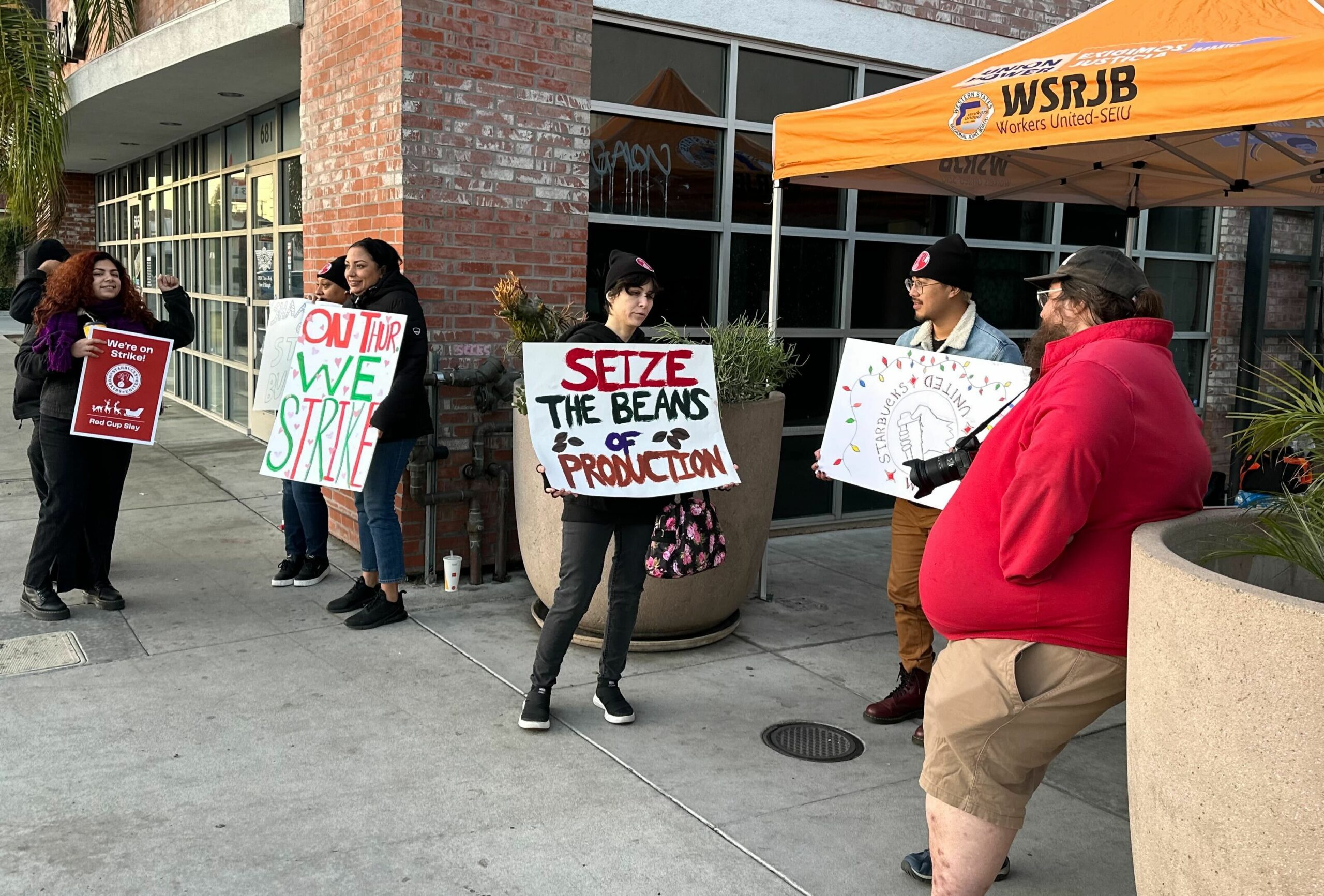Workers hold signs that read, “Starbucks Workers United,” “ON THUR[S] WE STRIKE,” and “SEIZE THE BEANS OF PRODUCTION,” with drawings of Christmas lights and coffee beans.