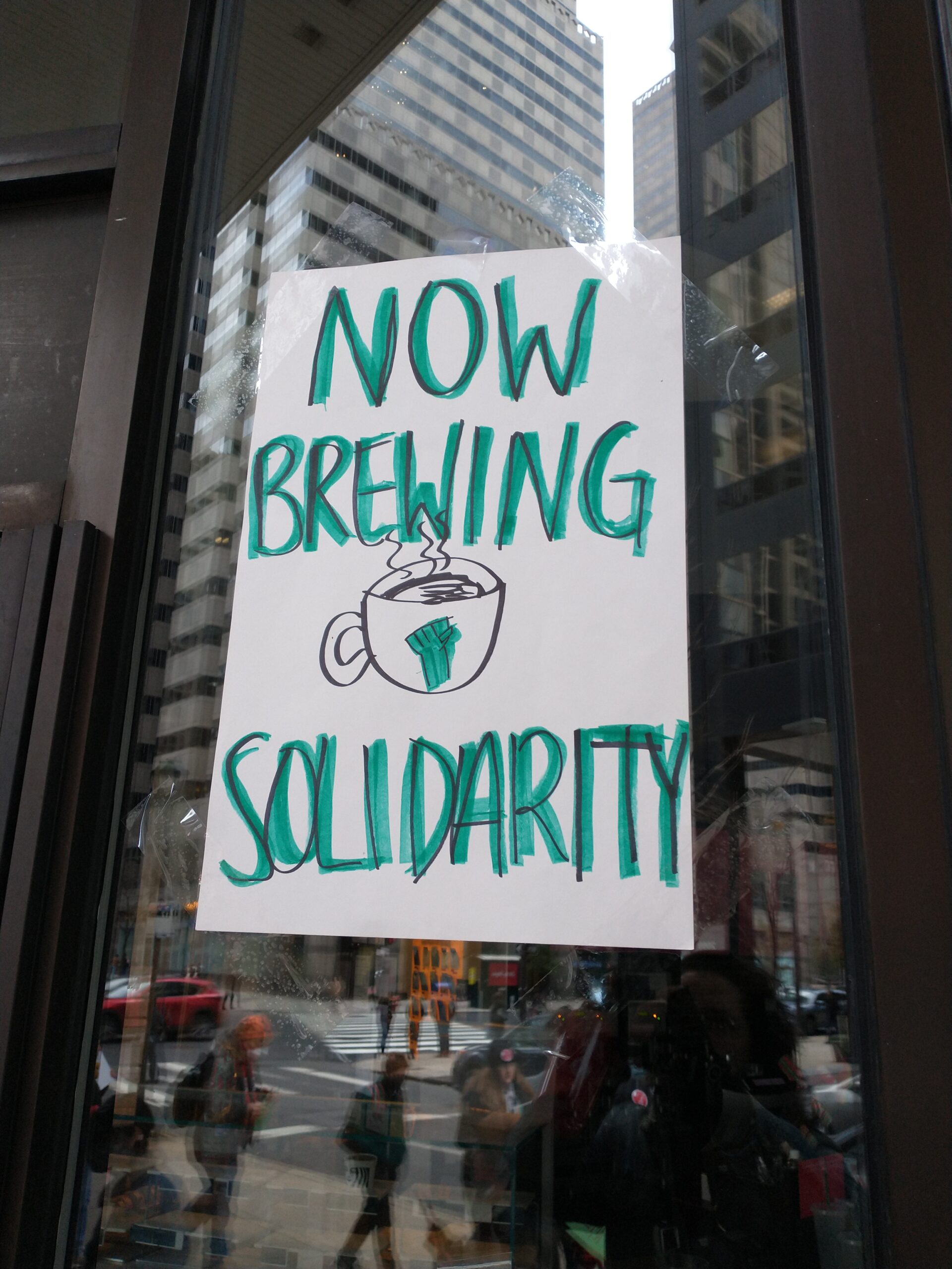 A hand-drawn sign taped to the glass window of a Starbucks store, written in the same green as the Starbucks logo, reads, “NOW BREWING SOLIDARITY.” Above the word “SOLIDARITY” is a drawing of a steaming cup of coffee with a green solidarity fist.