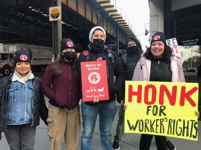 Five Starbucks workers pose for the camera wearing black hats with red Starbucks Workers United logo. Two workers hold signs: one reads, “We’re on Strike! Red Cup Slay” with the same union logo and the silhouette of Santa’s sleigh being led by reindeer; the second reads, “HONK FOR WORKERS’ RIGHTS.”