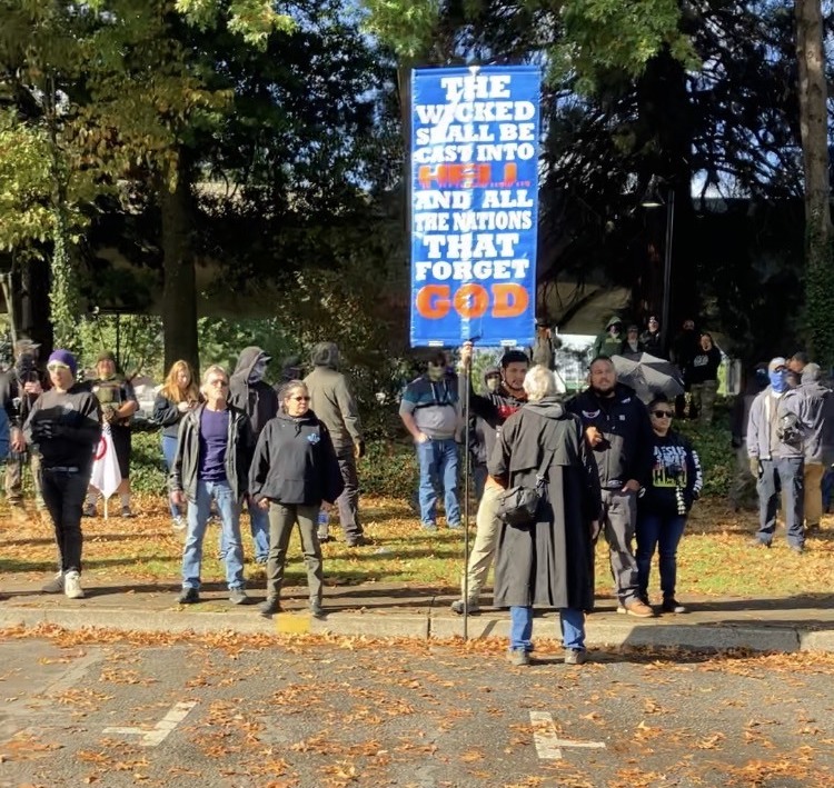 A crowd of protesters stand along a sidewalk and in the grass behind them. In the center is a man holding a large blue vertical banner on a poll that reads, “THE WICKED SHALL BE CAST INTO HELL AND ALL THE NATIONS THAT FORGET GOD,” with the words “HELL” and “GOD” written in red.