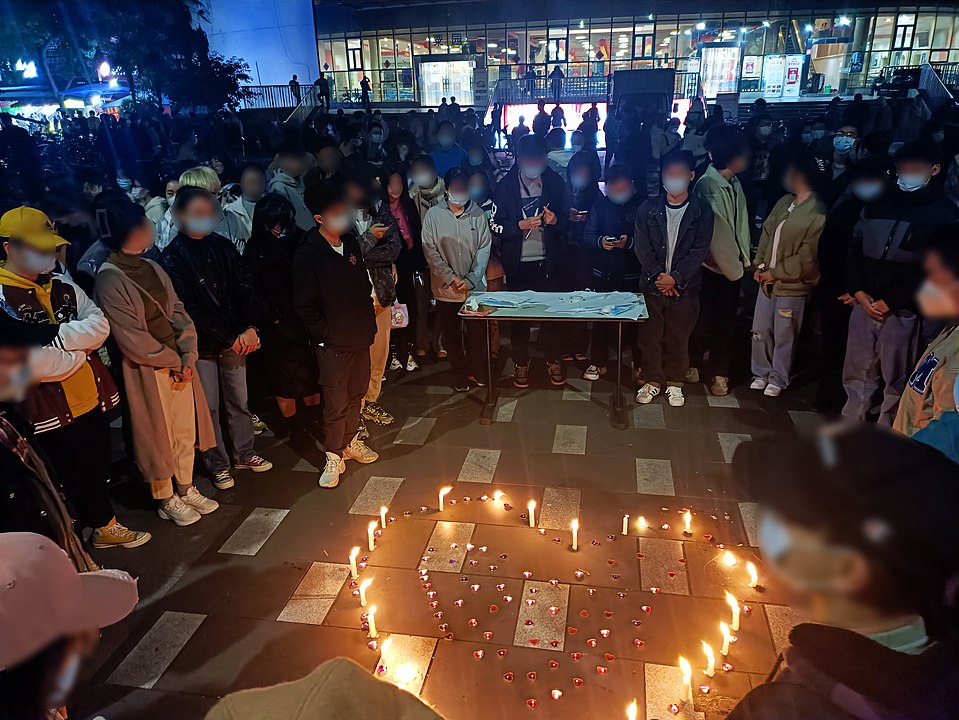 A large group of students of Southwest Jiotong University stand outside a university hall, in front of a table, and surrounding a makeshift altar, made out of  burning candles in the shape of a heart, to mourn the victims of the Urumqi fire. 