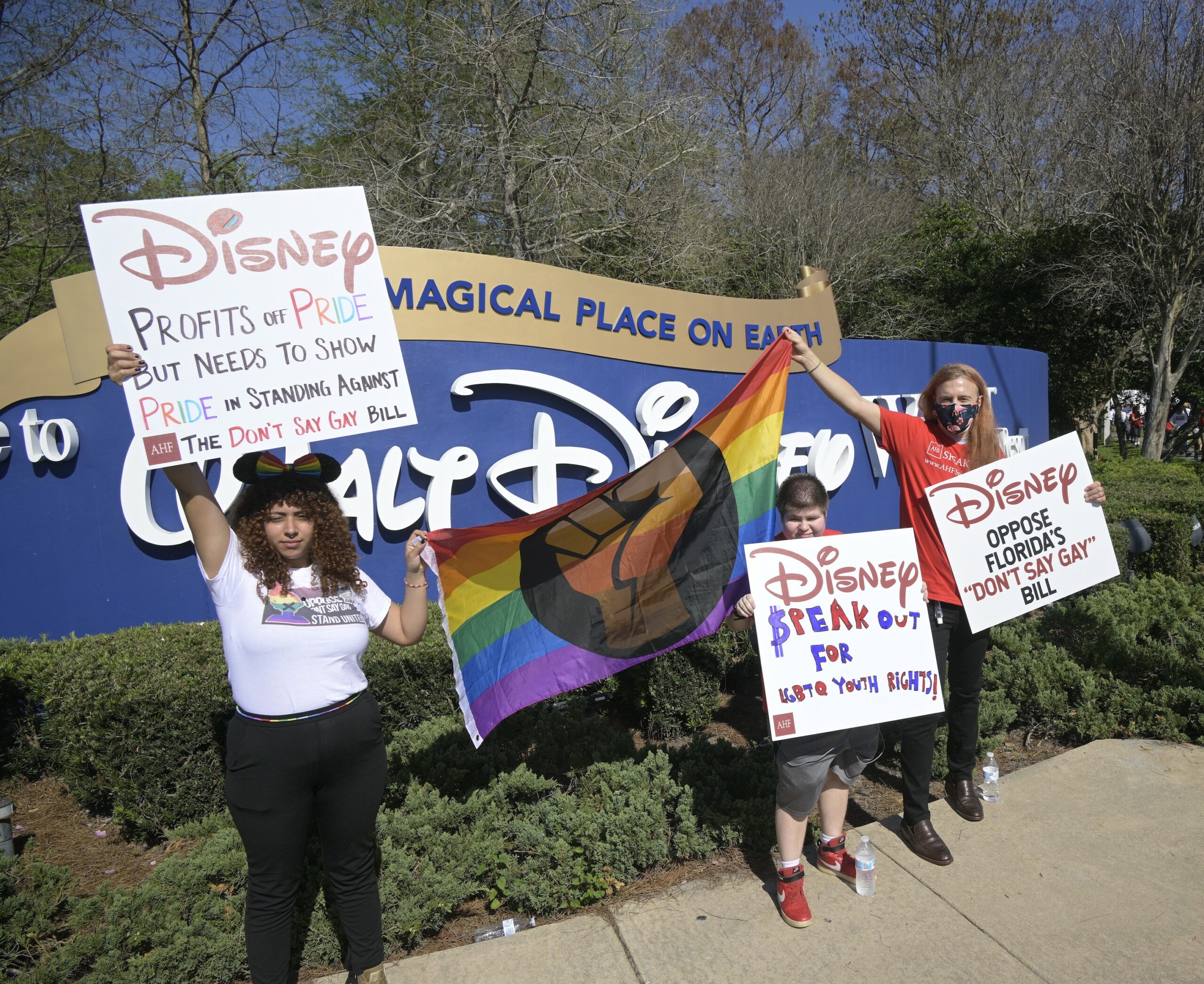 hree people stand in front of the “Welcome to Walt Disney World” theme park holding protest signs. Two of the people hold opposite ends of an LGBTQ pride flag with a brown fist in the center. Their signs read, “DISNEY PROFITS OFF PRIDE BUT NEEDS TO SHOW PRIDE IN STANDING AGAINST THE DON’T SAY GAY BILL;” “DISNEY SPEAK OUT FOR LGBTQ YOUTH RIGHTS;” and “DISNEY OPPOSE FLORIDA’S ‘DON’T SAY GAY’ BILL.”