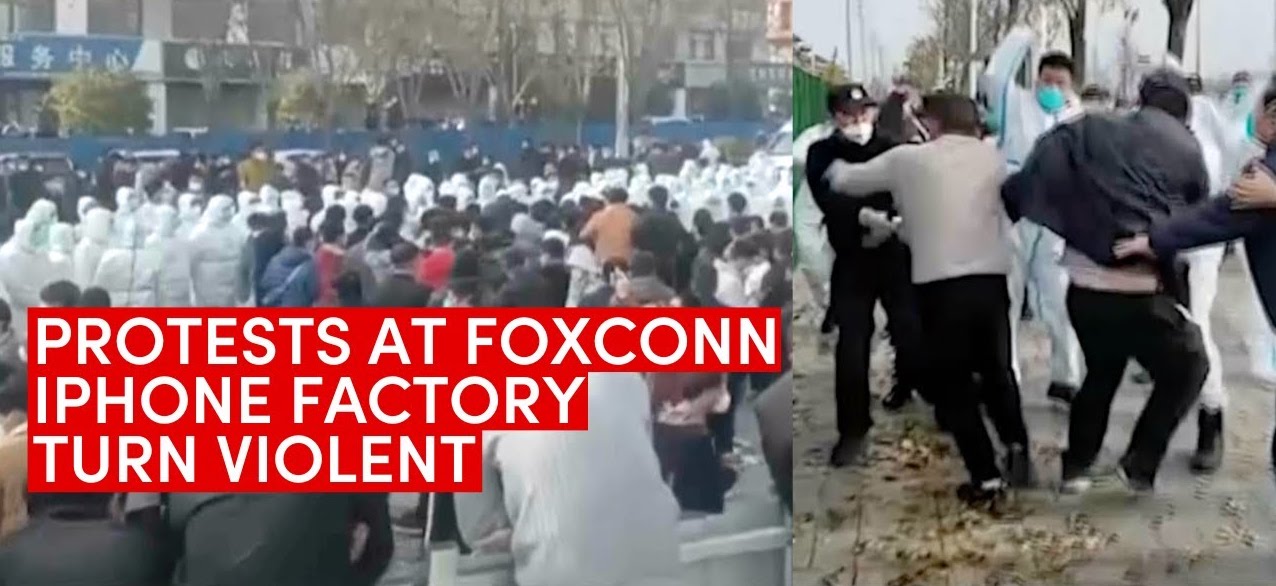 Screen shot of workers from the Foxconn Apple manufacturing plant in Zhengzhou, China, many in white boilersuits and masks, fighting with police and plant management during the recent industrial uprising November 23, 2022 with the large caption reading: “Protests at Foxconn iPhone factory turn violent.”