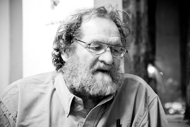 A black and white image of British Marxist John Molyneux. He is a large man with a gray beard and wire-rimmed classes and is wearing a button-down Oxford shirt.