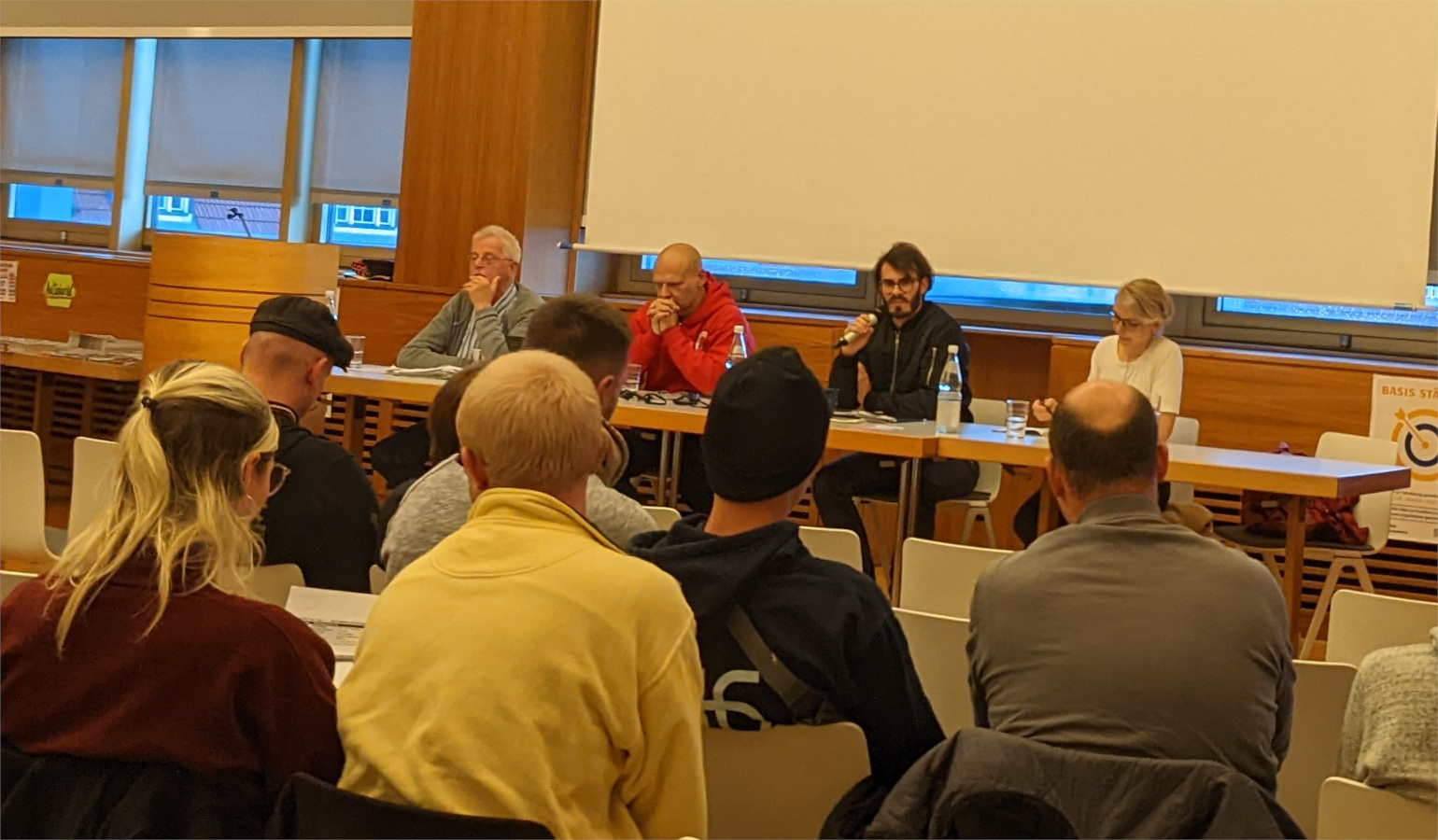 Photograph from the Organisieren, Kämpfen, Gewinnen conference in Germany, September, 2022, which shows four speakers at a table at the front of the room speaking to people (nine shown), with their backs to the photographer.