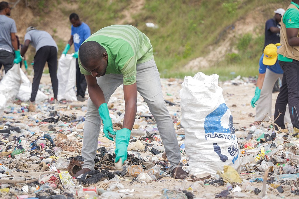 A Black man is front and center wearing a green shirt, gray pants, brown shoes, and green protective gloves is picking up a piece of plastic to dispose of in a large white bag. Their are small groups of individuals on each side of the picture doing the same also wearing the green protective gloves. The beach is in Ghanna and is covered in a number of different kinds of plastic waste.