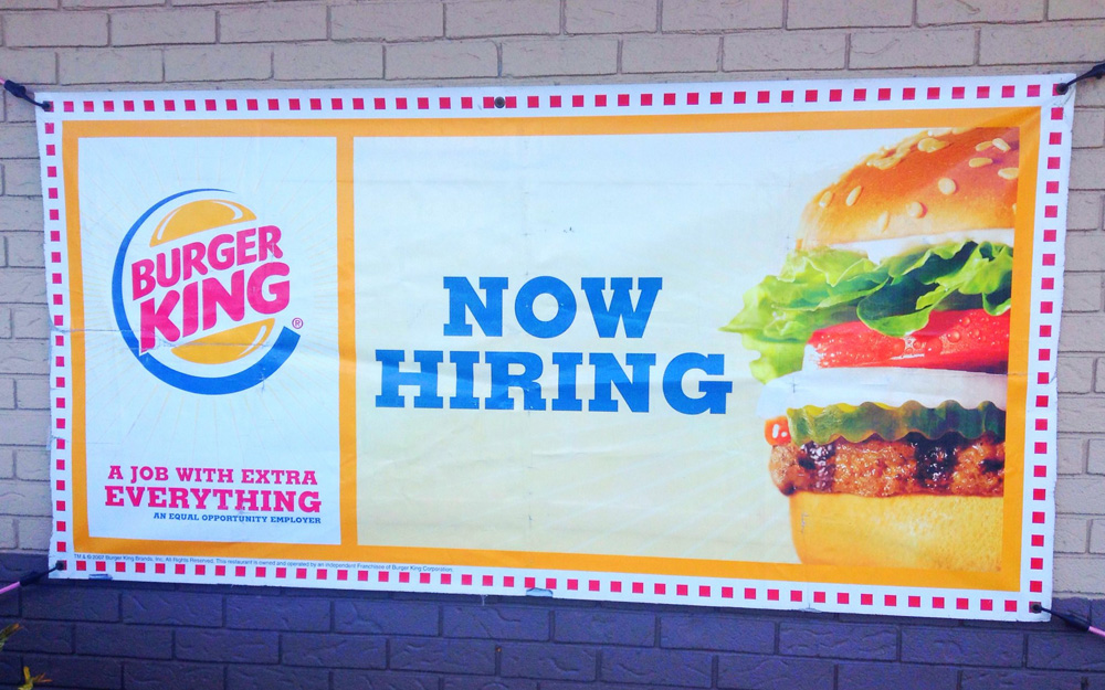 A Burger King help wanted sign reading "Now Hiring" with an image of a hamburger.