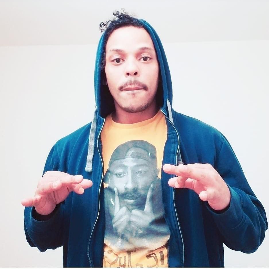 Nzoy poses for a picture with his hands slightly raised in a blue hoodie and a yellow shirt with a large image of U.S.-born rapper Tupac Shakur (also known as 2Pac and Makaveli) on the shirt against a white backdrop.