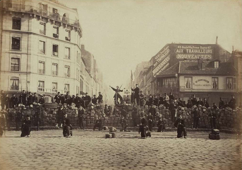 A vintage black and white photograph of workers in black building a barricade in the streets of Paris in 1871. 