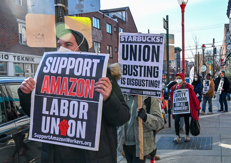 A line of picketers carry signs reading "Support Amazon Labor Union" and "Starbucks Union Busting is Disgusting."