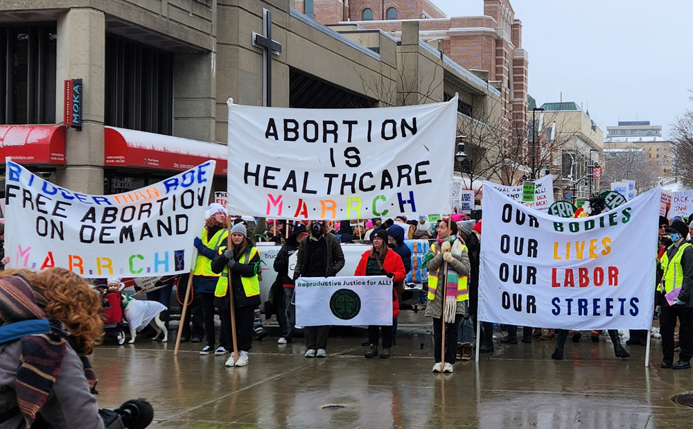 Activists line up on a rainy street in downtown Madison, Wisconsin carrying bright banners that read Bigger Than Roe: Free Abortion on Demand, Abortion is Healthcare, and Our Bodies, Our Lives, Our Labor, Our Streets. The name of the organization behind the event, M.A.R.R.C.H., is on teh banners. There is a small banner reading "Reproductive Justice for All."