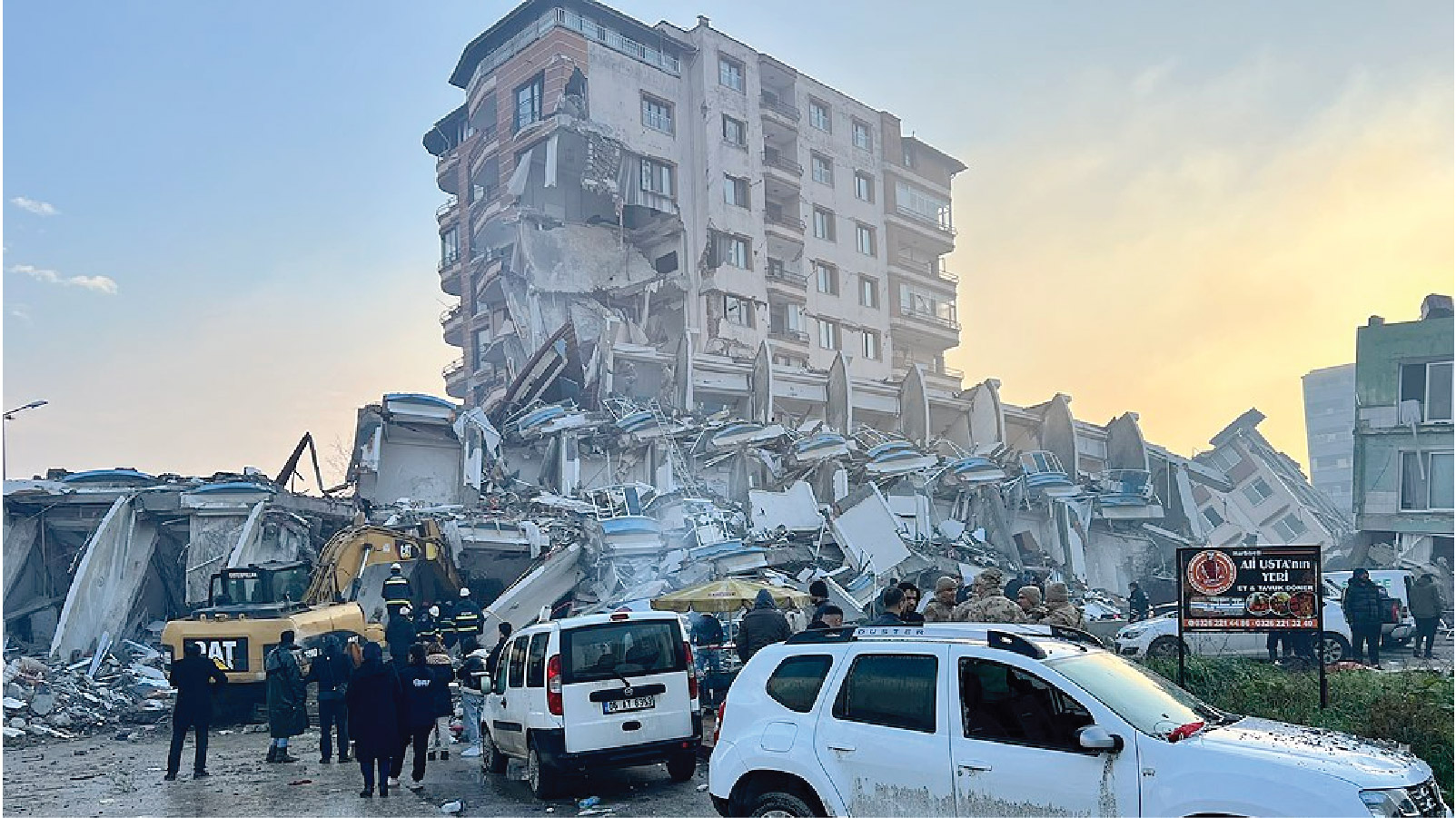 A block of destroyed buildings in the Hatay province of Türkiye shows the earthquake’s significant impact. One building is still standing but has significant damage and looks like it is on the brink of collapse. It is surrounded by rubble of collapsed buildings. An excavator works on the rubble as people observe the scene. Parked vehicles are in the forefront.