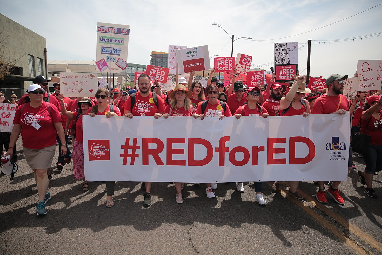People wearing red t-shirts that say ‘Arizona Educators United’ march down a street with various signs and a banner that says ‘#REDforED.’ One sign says ‘Education Cuts NEVER Heal’ with band-aids and another says ‘Quality Ed Deserves Quality Pay.’