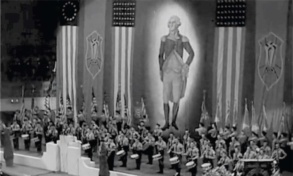A black-and-white photo of a large stage with a band ensemble of drummers and additional musicians, standing in front of multiple U.S. flags on stands. A large banner of George Washington descends from the atrium’s ceiling, along with large German American Bund flags and the Confederate States of America flag.