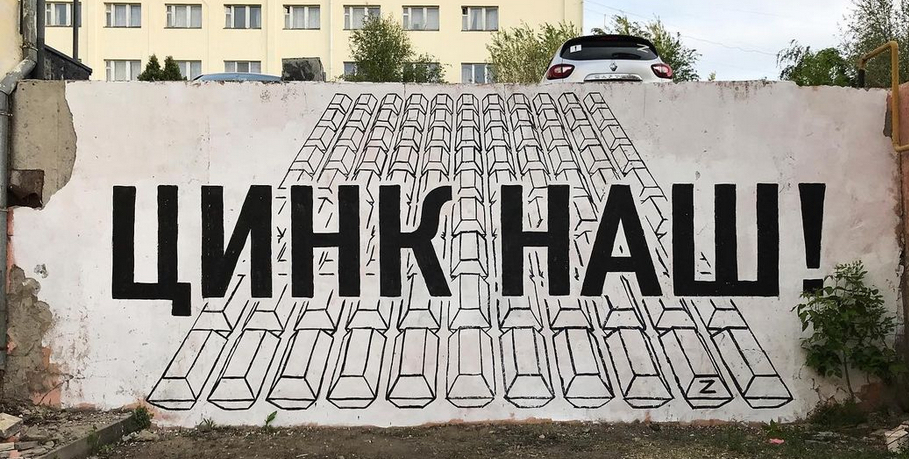 Russian words ‘цинк наш!’ graffitied on a white-painted outdoor wall about 12 ft tall. The letters are capitalized and are layered over graffitied images of coffins. There is a small graffiti signature of ‘Z.’ The forefront ground is dirt with some grass. The background shows a car, an apartment building and trees.