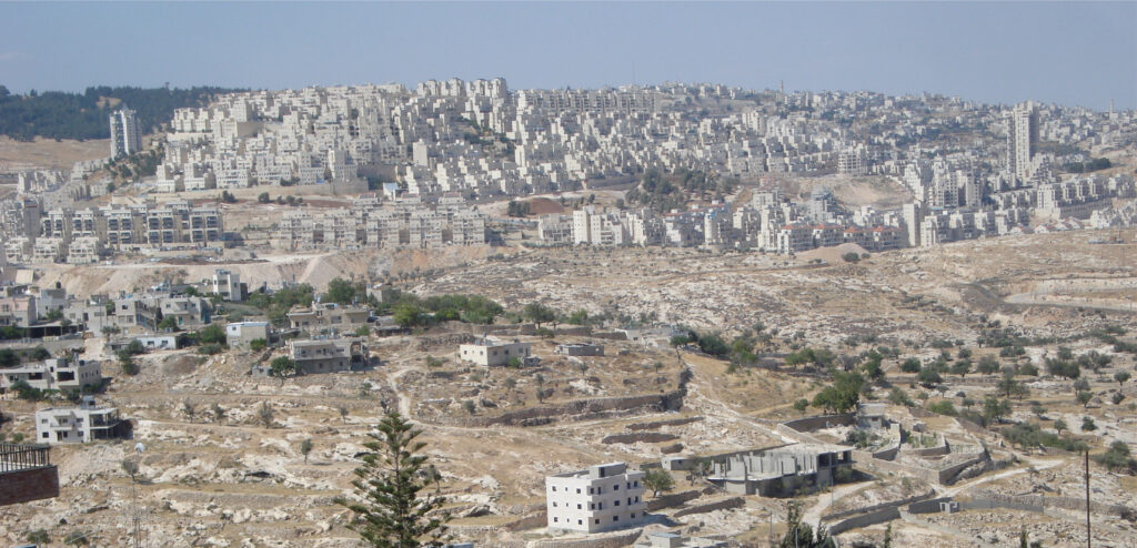 In the top half of the photo, a large and concentrated amount of white apartment buildings engulf the top half of a tall and wide hill in the photo’s view. Part of the large hill below the buildings is undeveloped and rocky land. In the forefront, and lower half of the photo, a smaller hill is majority undeveloped land, composed of brownish-yellowish rocky terrain with a bit of grass and speckled with a couple of one to three floor buildings. It looks like there are layers in the hill which are most likely roads paved into the hillside. Some trees speckle the undeveloped parts of both hills.