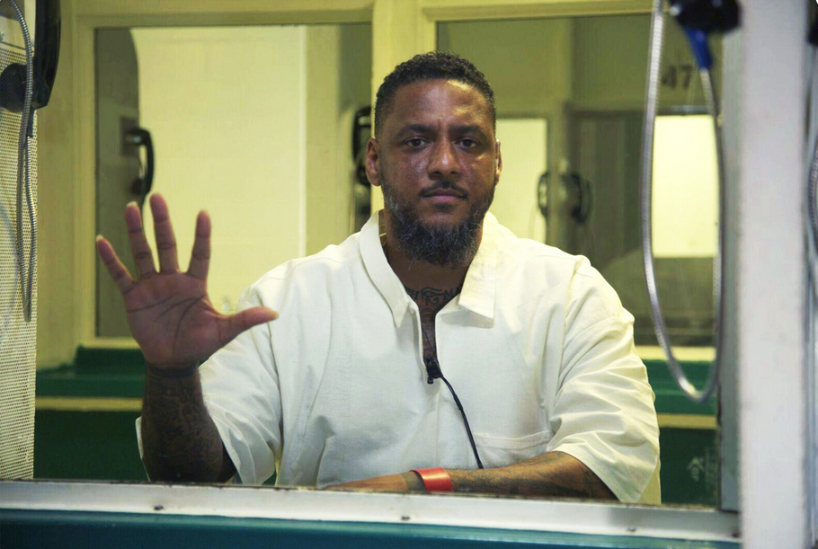 Kenneth Foster Jr. sits in a prison visitation booth, looks straight on at the camera and holds his hand up with his palm against the glass that separates visitors from prisoners. Surrounding mirrors show other visitation booths with their separation glass and phones.