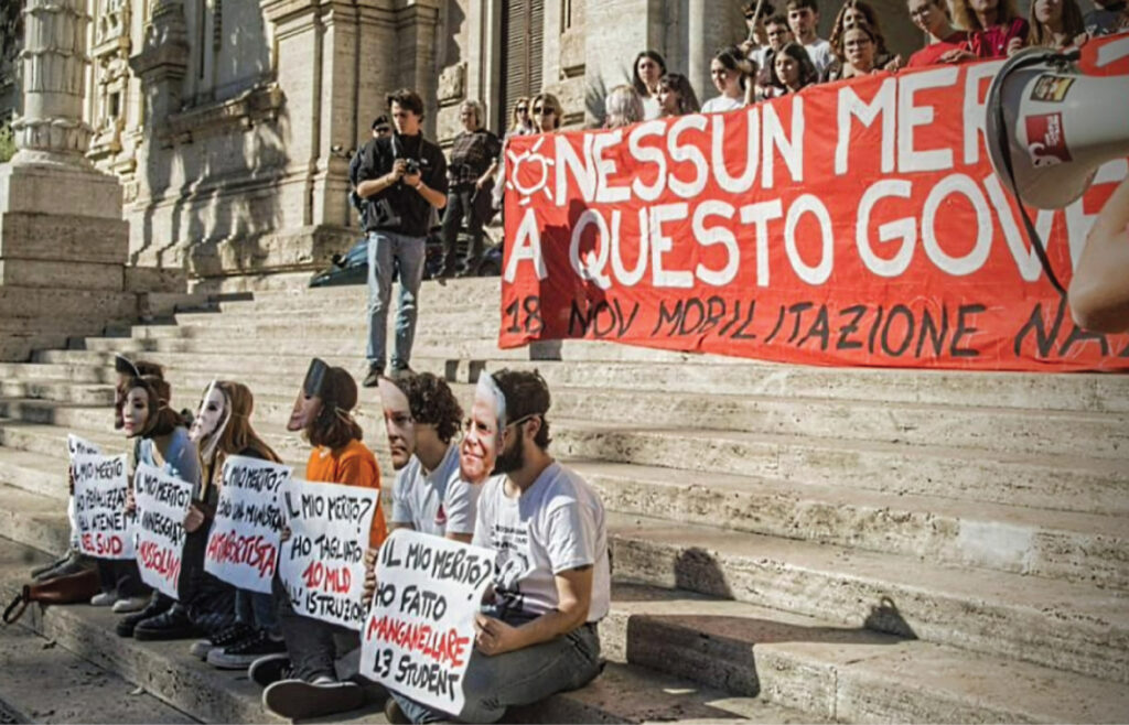 Alt Text for Image: In the forefront, a group of six younger people sit on steps of a Romanesque building, wearing masks of faces of the recently elected Italian government and holding up various signs, each beginning with “IL MIO MERITO?” In the background, more people hold up a large red banner with text partly covered by someone using a megaphone. The text showing says “NESSUN MER… A QUESTO GOV… 18 NOV MOBILITAZIONE …”. A photographer walks amid the demonstration.