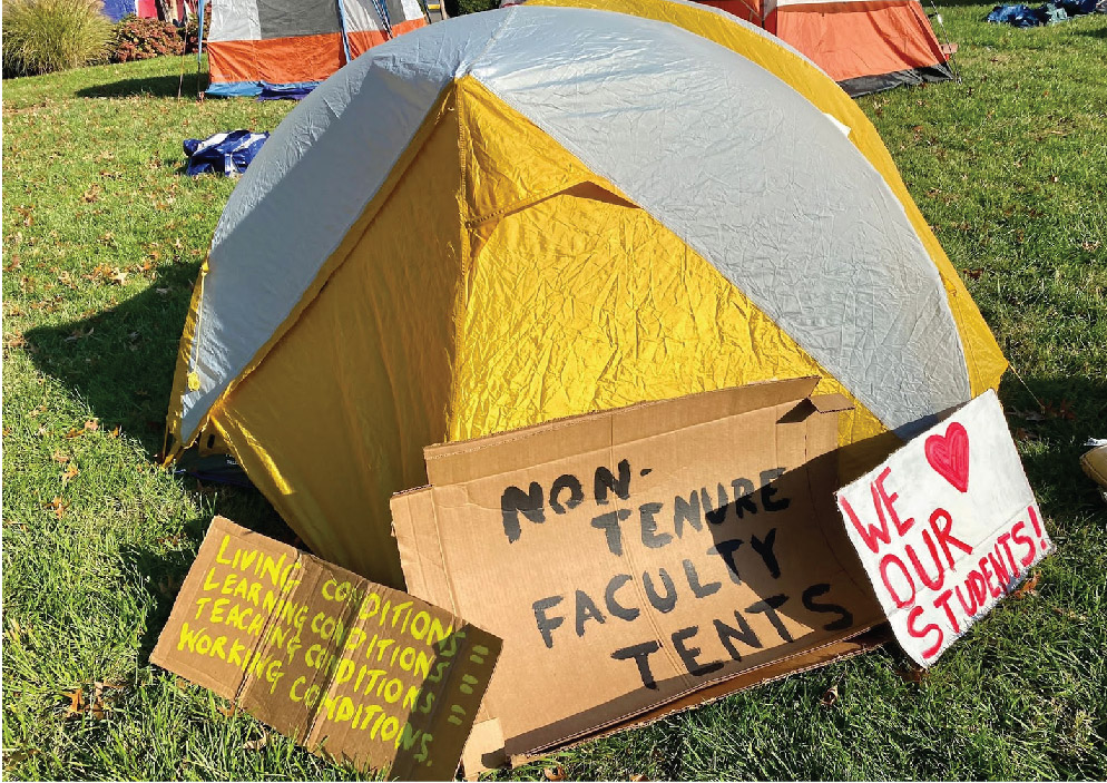 In the forefront, a yellow and beige tent stands with various cardboard signs leaned against it. One sign reads “LIVING CONDITIONS = LEARNING CONDITIONS = TEACHING CONDITIONS = WORKING CONDITIONS.” Another reads “NON-TENURE FACULTY TENTS.” And the last reads “WE LOVE OUR STUDENTS!”. Several orange tents are in the background.