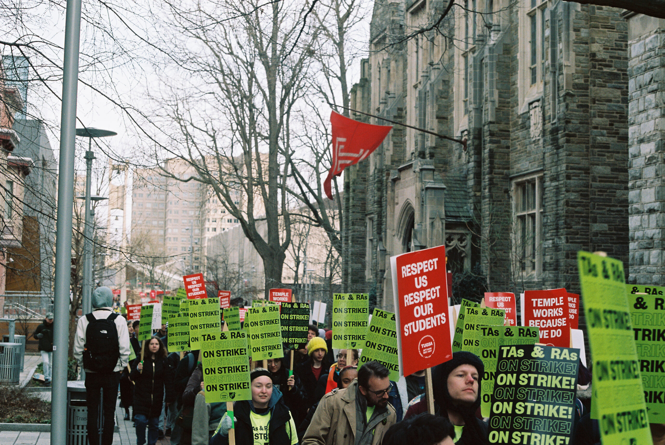 A large group of Temple graduate workers is bundled up in winter clothes while they are on strike, marching together through the university's main campus outside of a historic building in North Philadelphia. The building features prominently a red Temple flag with a white T outlined on the flag. Most are carrying signs, many of the signs read "TAs & RAs ON STRIKE!" repeated multiple times in a green background with black text, and others have red signs, some of them read "RESPECT US RESPECT OUR STUDENTS" and others have the message "TEMPLE WORKS BECAUSE WE DO."