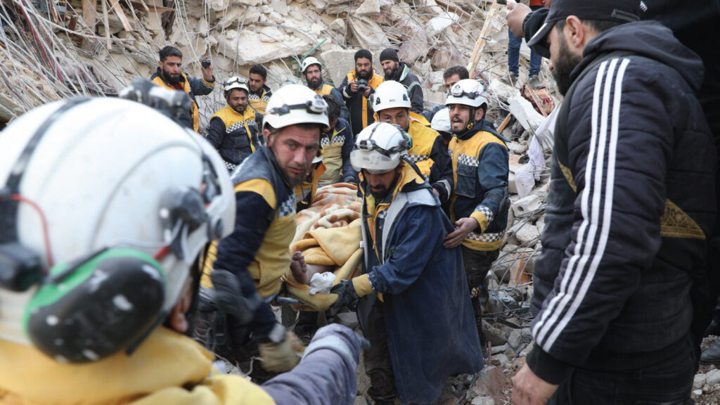 A group of The White Helmets volunteers engage in a rescue effort in Northwest Syria on February 8, 2023. Several volunteers carry a stretcher with an injured individual away from a destroyed building, surrounded by rubble. More volunteers and observers stand nearby, watching or taking photos of the rescue effort.