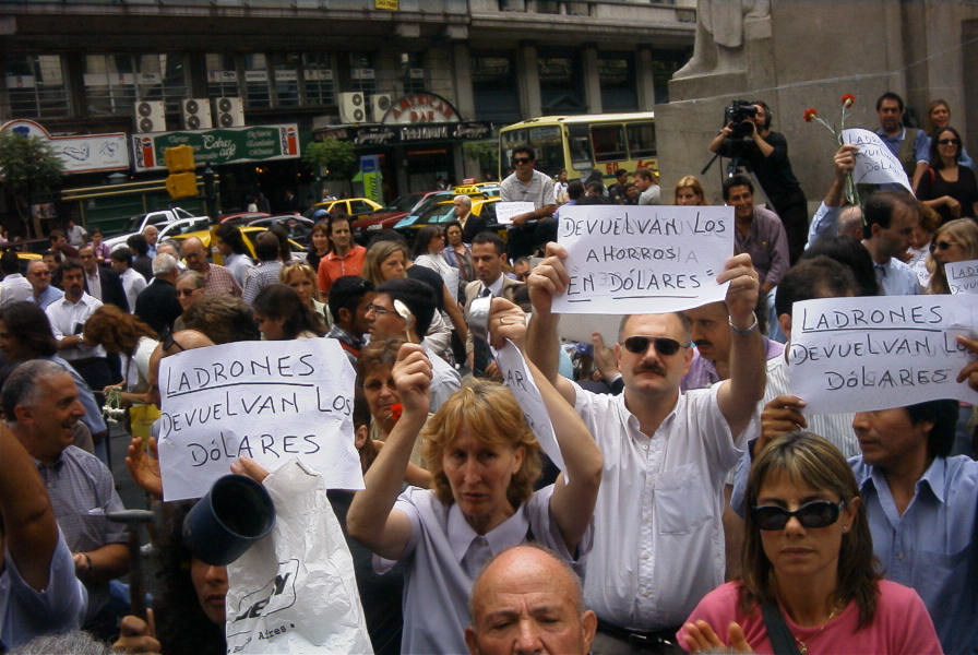 Many people occupy an outdoor area to protest a bank they are standing in front of. They are holding up various signs, flowers, and dishware to use to make sound - for example, one person is hitting a spoon against a little metal cup. Two signs read ‘LADRONES - DEVUELVAN LOS DÓLARES’ which in English roughly means, THIEVES - RETURN THE DOLLARS. A third sign reads ‘DEVUELVAN LOS AHORROS EN DOLARES =’ which in English roughly means, RETURN THE SAVINGS IN DOLLARS. A cameraperson stands in the background, filming the scene. Storefronts and a street with various cars, taxis and a bus, are in the background.