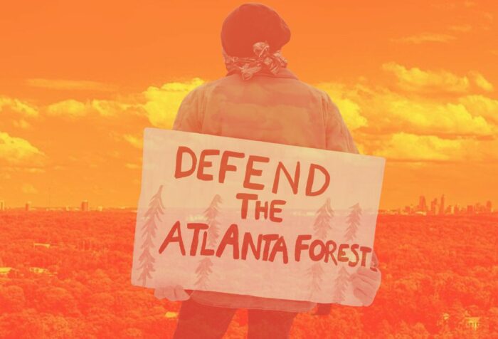 A protester on a yellow background stands in a field holding a sign reading DEFEND THE ATLANTA FOREST.