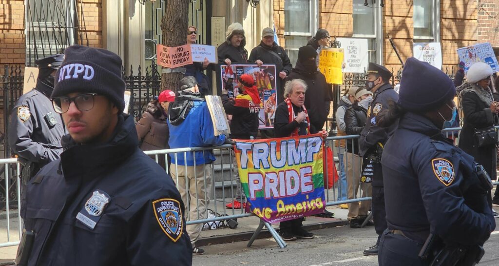 A photo of the protesters against the Drag Story Hour standing on a sidewalk, hemmed in by mobile metal fences placed alongside the sidewalk on the street. One person is wearing a ‘gay pride flag’ pin on their shirt and holds up a flag with the rainbow coloring of the gay pride flag with text that says ‘TRUMP PRIDE Save America.’ About 10 other protesters stand behind the fence, holding various transphobic and homophobic signs. One sign reads ‘Stop Taking Children’s Innocence Away.’ Several NYPD cops stand along this fence as well as in the forefront of the photo, looking in various directions and not engaged with any of the protesters.