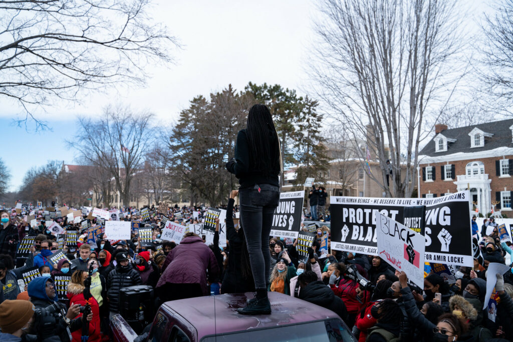 A person stands on the top of a truck and, using a microphone and speaker system in the truck’s trunk, addresses a large crowd of people protesting and occupying a residential street. About half of the protesters are holding signs, most that read as ‘PROTEST YOUNG BLACK LIVES’, another says ‘BLACK LIVES ALWAY SMATTER’ with a Black power fist, another reads ‘JUSTICE FOR AMIR LOCKE.’ Others hold up three large banners that read ‘WE ARE HUMAN BEINGS - WE DESERVE TO LIVE - WE HAVE HOPES & DREAMS - PROTECT YOUNG BLACK LIVES - JUSTICE FOR AMIR LOCKE’ and have two power fists. Most people are wearing hats and winter coats.