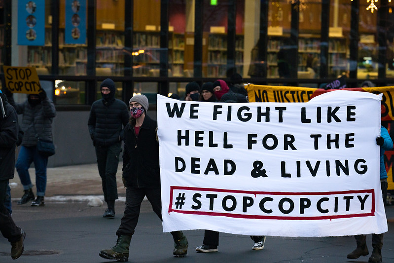About 10 people are walking down a street as part of a larger march. One person carries a sign that reads ‘STOP COP CITY.’ Two people hold up a large banner, as tall or taller than them, that reads ‘WE FIGHT LIKE HELL FOR THE DEAD & LIVING - #STOPCOPCITY.’ Another large banner is behind the first one, but most of its language is cut off - the viewer can only see the words ‘NO JUSTICE…’ All are wearing hats and coats in the chilly weather, and you can see some snow along the sidewalk. A lit up storefront, or library, with many shelves of books, stands in the background.