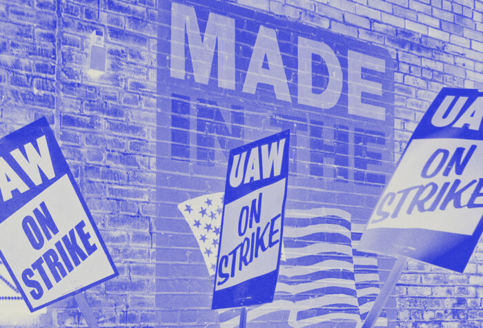 Three signs in blue on white read UAW on strike in front of a sign reading MADE IN THE [American flag}.
